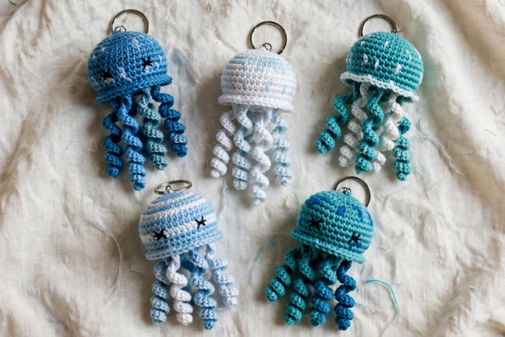 Five blue, teal and white crochet jellyfish keychains with curly tentacles arranged on pale linen cloth